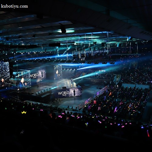 A large indoor arena filled with an audience watching a live performance. The stage features large screens displaying a performer and graphics, with bright spotlights and ambient lighting creating a dynamic atmosphere. The crowd is holding glow sticks, adding vibrant colors to the scene.