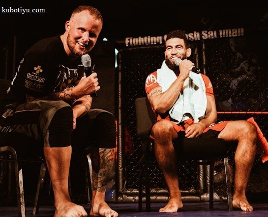 Two individuals are seated on a stage, each holding a microphone and smiling. They are in a sports-related setting with one wearing a casual black outfit and the other in sportswear, with a towel around his neck. The background includes elements of a fighting or martial arts cage.