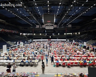 An indoor sports arena filled with rows of tables, each occupied by people engaged in competitive activities. The ceiling is high, with numerous lights illuminating the large space. The audience seating on either side is empty, and there are flags hanging from the upper level.