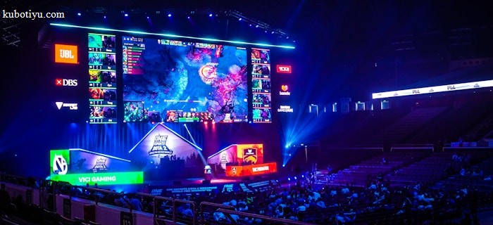A large esports arena featuring a massive screen displaying a video game, surrounded by sponsor logos and team names. The auditorium is filled with spectators watching the gaming event. Neon blue and purple lights illuminate the stage, creating a vibrant atmosphere.