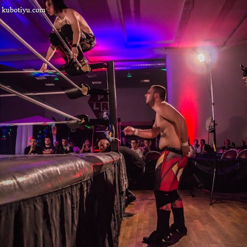 A wrestling match taking place in an indoor venue, where one participant is crouched on the top rope of the ring, while the other stands outside the ring looking at them. A crowd watches the event enthusiastically from their seats.