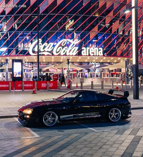 A sleek black sports car is parked in front of an illuminated arena entrance, with a large sign bearing the name of a famous beverage brand. The building features a modern, geometric facade with bright red and blue lights, creating a vibrant and lively atmosphere.